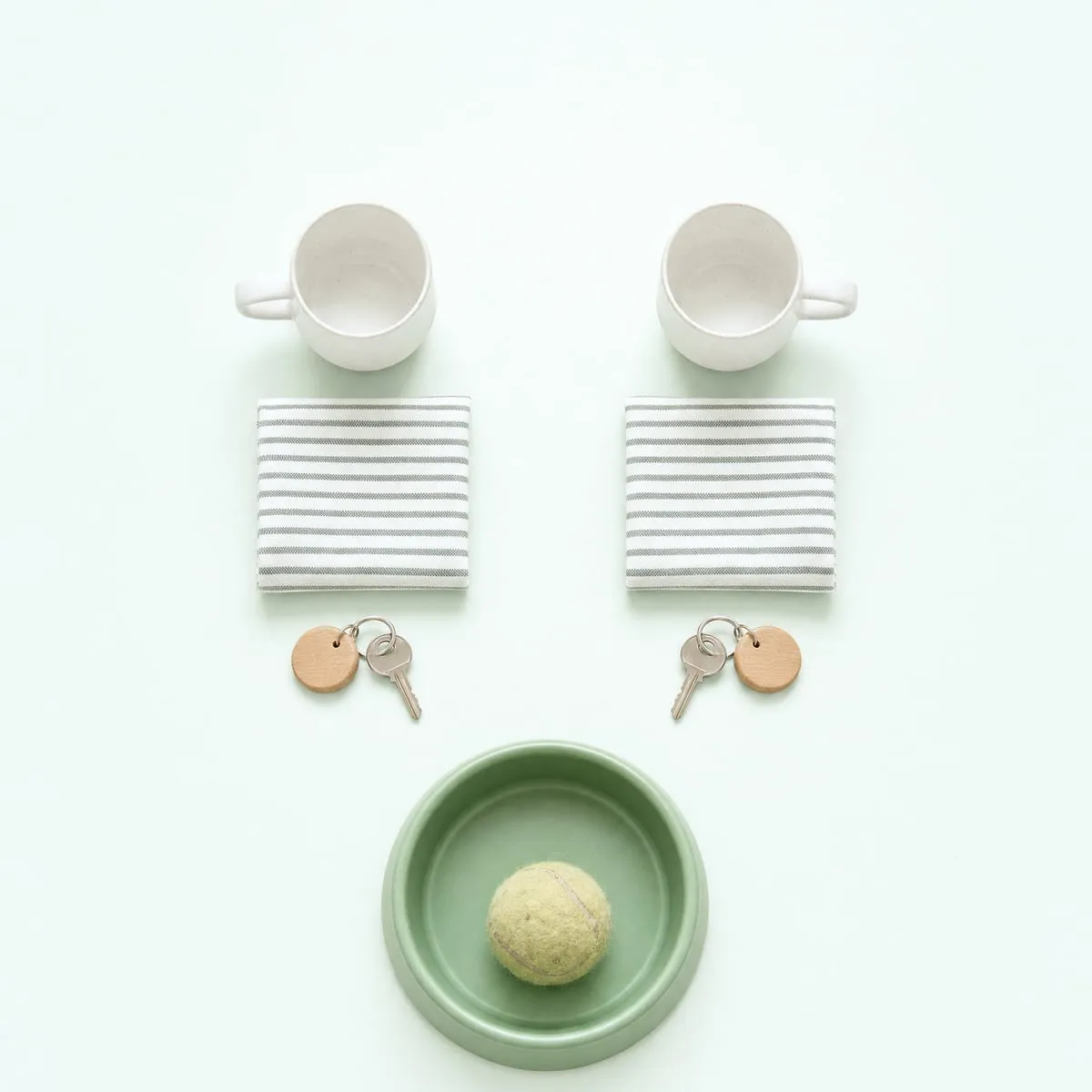 Two mugs, two folded tea towels and two sets of keys laid out in matching pairs, with a single dog bowl below them.